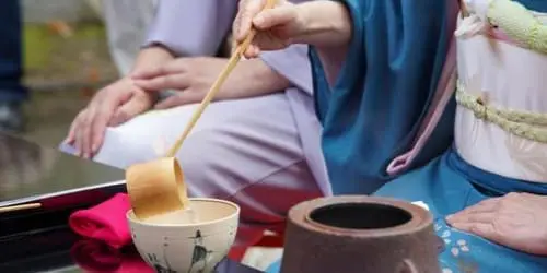 民族衣装を着た人物が竹の柄杓を使って、装飾された椀に液体を注いでいる。近くの台には陶器の壺とピンク色の布が置かれている。背景には別の人物が座っている。.