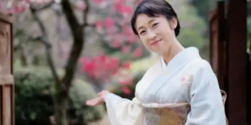 淡い色の着物を着た人が、木造建築物の近くの屋外に立って、片手で合図している。背景にはピンクの花が咲いている。.