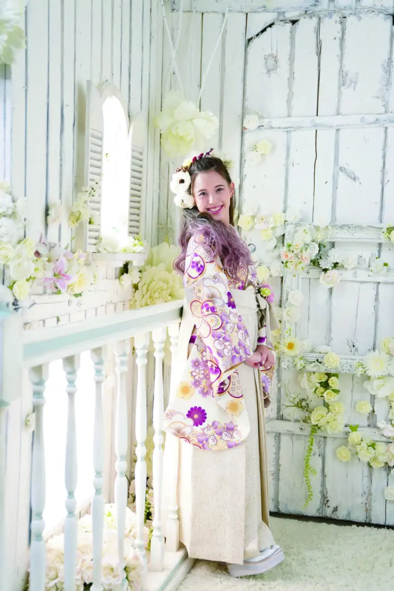 素朴な壁と明るい花柄で飾られた部屋で、白と紫の花柄の着物「レンタル袴 01」を着た人が白い手すりに寄りかかりながらカメラに向かって微笑んでいます。.