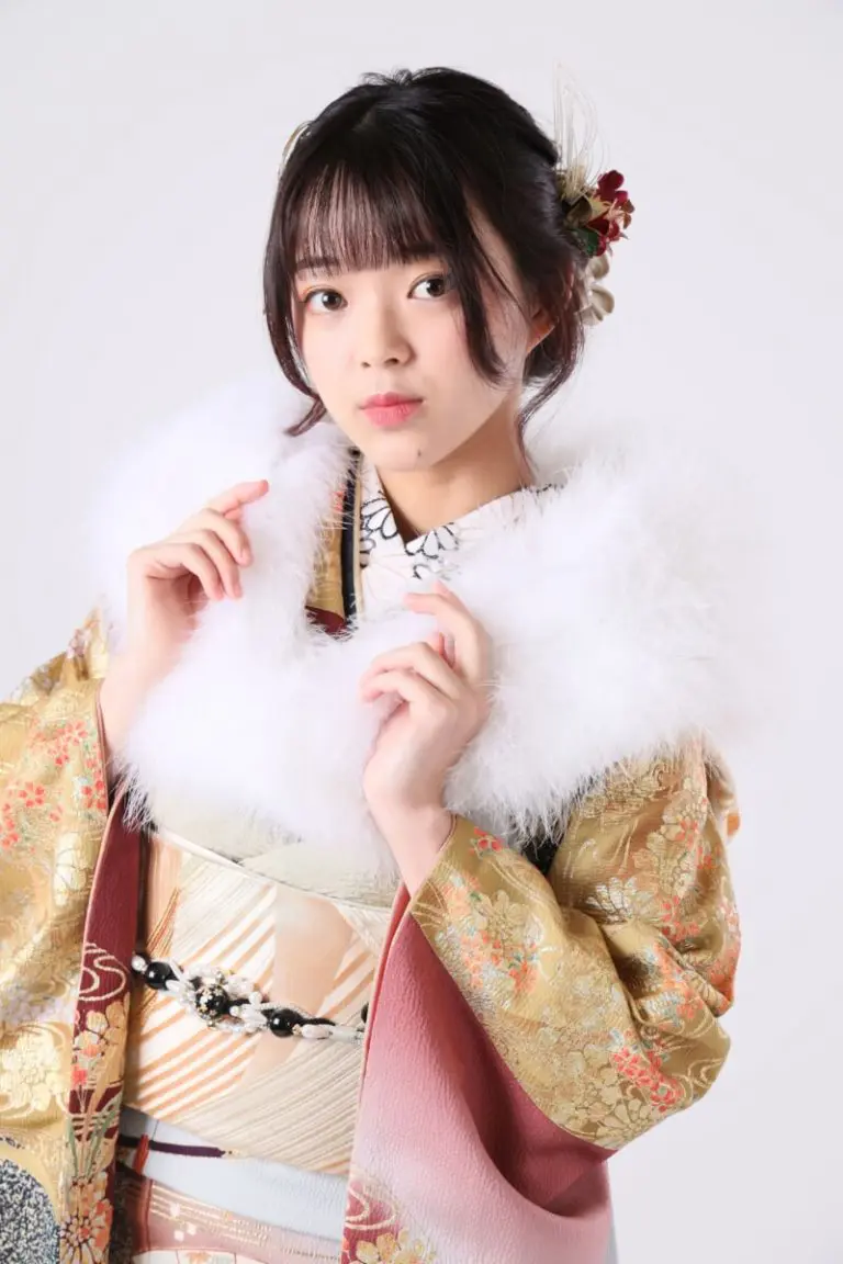人物がシンプルな背景に立っています。花柄の伝統的な日本の着物を着て、白いふわふわのショールを肩にかけています。黒髪には花飾りが施されています。.