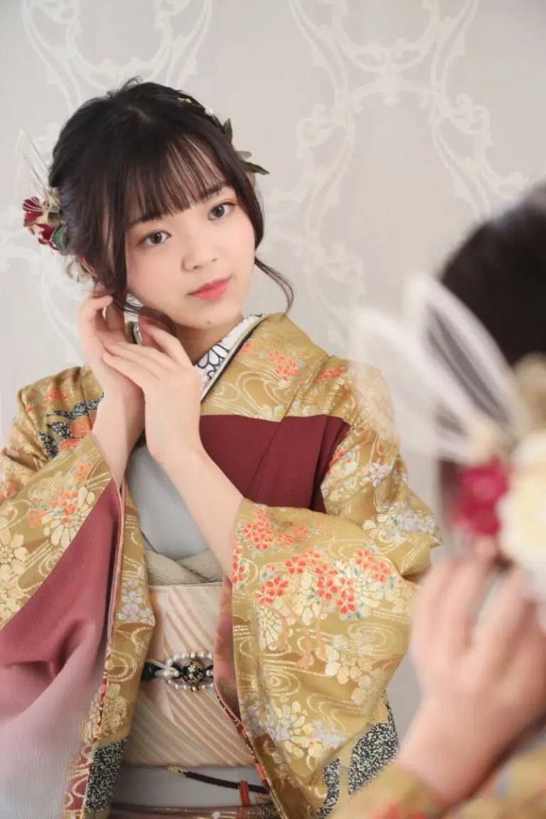 花柄の着物を着た女性が、鏡を見つめながらイヤリングを直している。着物には金と赤の模様が描かれ、髪型にも花飾りが添えられている。背景には、控えめな装飾の壁紙が使われている。.