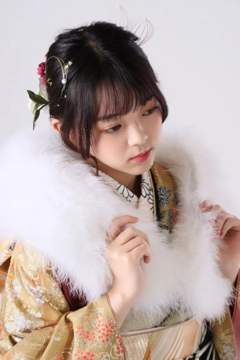 花と金の模様が入った伝統的な着物に白い毛皮の襟をつけた人物。髪には花やアクセサリーが飾られ、無地の背景を背に横を向いている。.