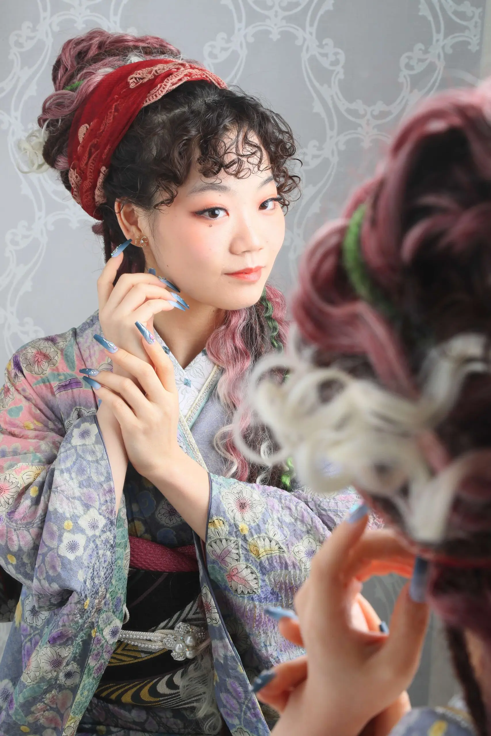赤いスカーフを巻いた巻き毛の人物が、繊細な模様の背景の前で、青とラベンダー色の花柄が描かれた戸田市の着物を着て、長く飾られた爪をしながら、鏡に映った自分の姿を見つめている。.