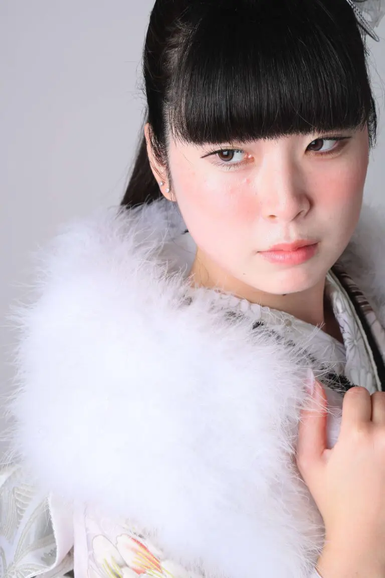 日本の伝統的な着物を着た女性が、白いふわふわのショールを肩にかけている。前髪をまとめ、物思いにふけるような表情を浮かべている。背景はライトグレー。.