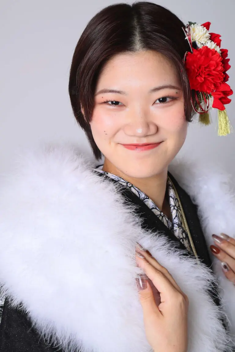 令和7年成人式、戸田市の伝統的な衣装を着た人物が、髪に赤と白の花を飾り、軽い毛皮を巻き、無地の背景を背景にカメラに向かって微笑んでいる。.