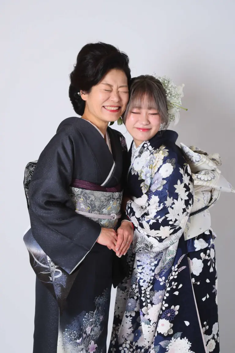 日本の伝統的な着物を着た二人の女性が、無地の白い背景に寄り添って微笑みながら立っています。左の女性は濃い色の着物を、右の女性は青い花柄の着物に花飾りを着けています。.