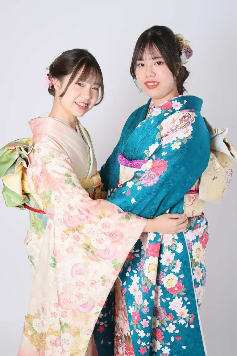 令和7年成人式、蕨市ラインの色鮮やかな着物を着た二人が並んで立っています。左は淡いピンクの花柄、右は青緑色の花柄の着物です。二人とも花のヘアピースを着け、カメラに向かって微笑んでいます。.