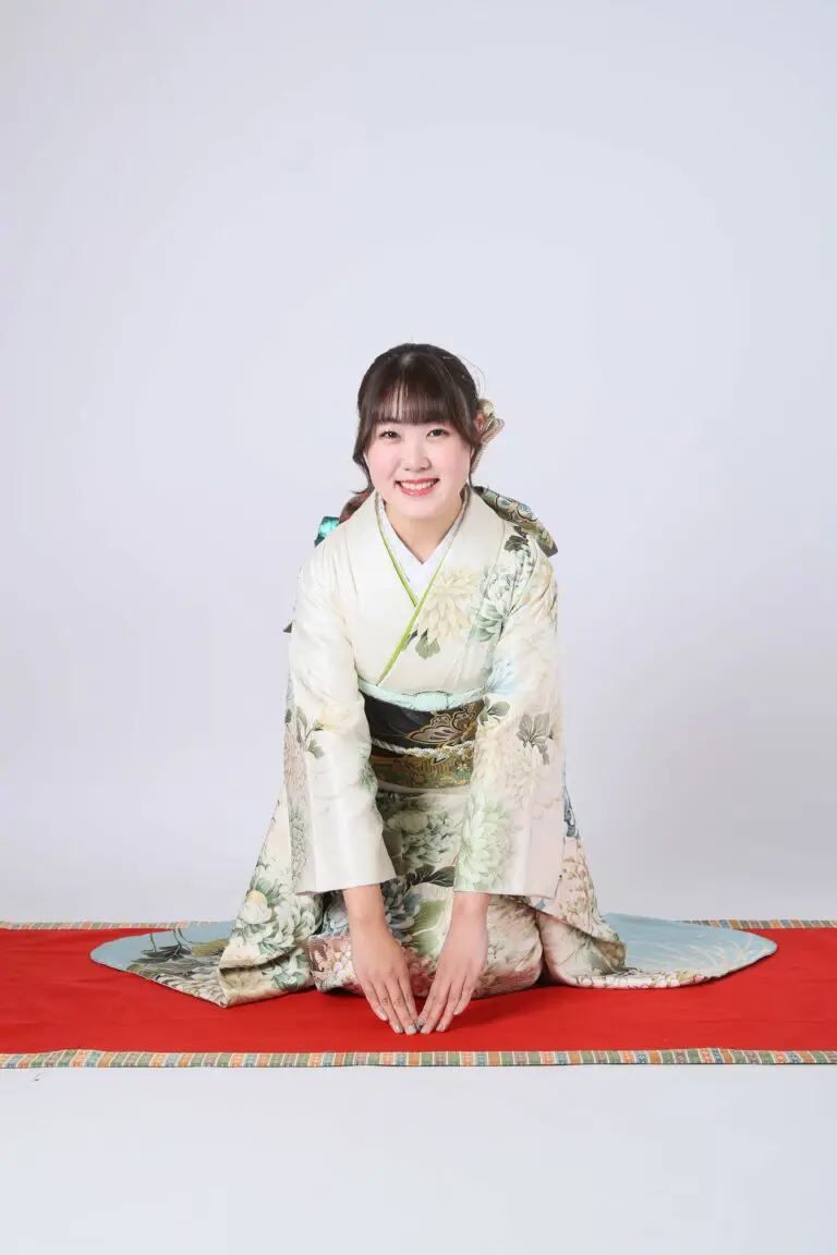 淡い色の花柄の着物を着た人物が、真っ白な背景の赤いマットの上に日本の伝統的な跪き姿勢で座っています。商品名：令和7年成人式、川口市。.