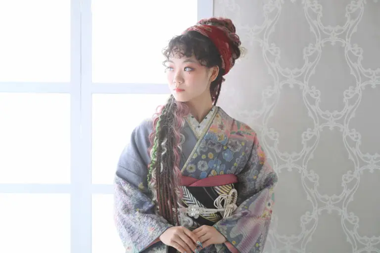令和7年成人式、戸田市コレクションの赤いかぶと柄の着物を着た人物が、室内で、複雑なデザインの壁のある柔らかな光が当たる窓際に立って、左を見つめている。.