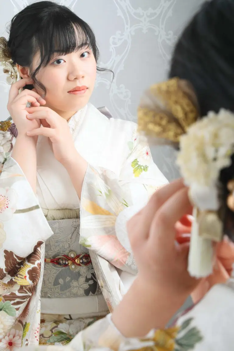 伝統的な花柄の着物を着た女性が、繊細な装飾模様を背景にした鏡を覗きながら髪飾りを調整しています。彼女は自分の反省に深く集中しています。このシーンには、令和7年成人式、川口市の商品が登場します。.