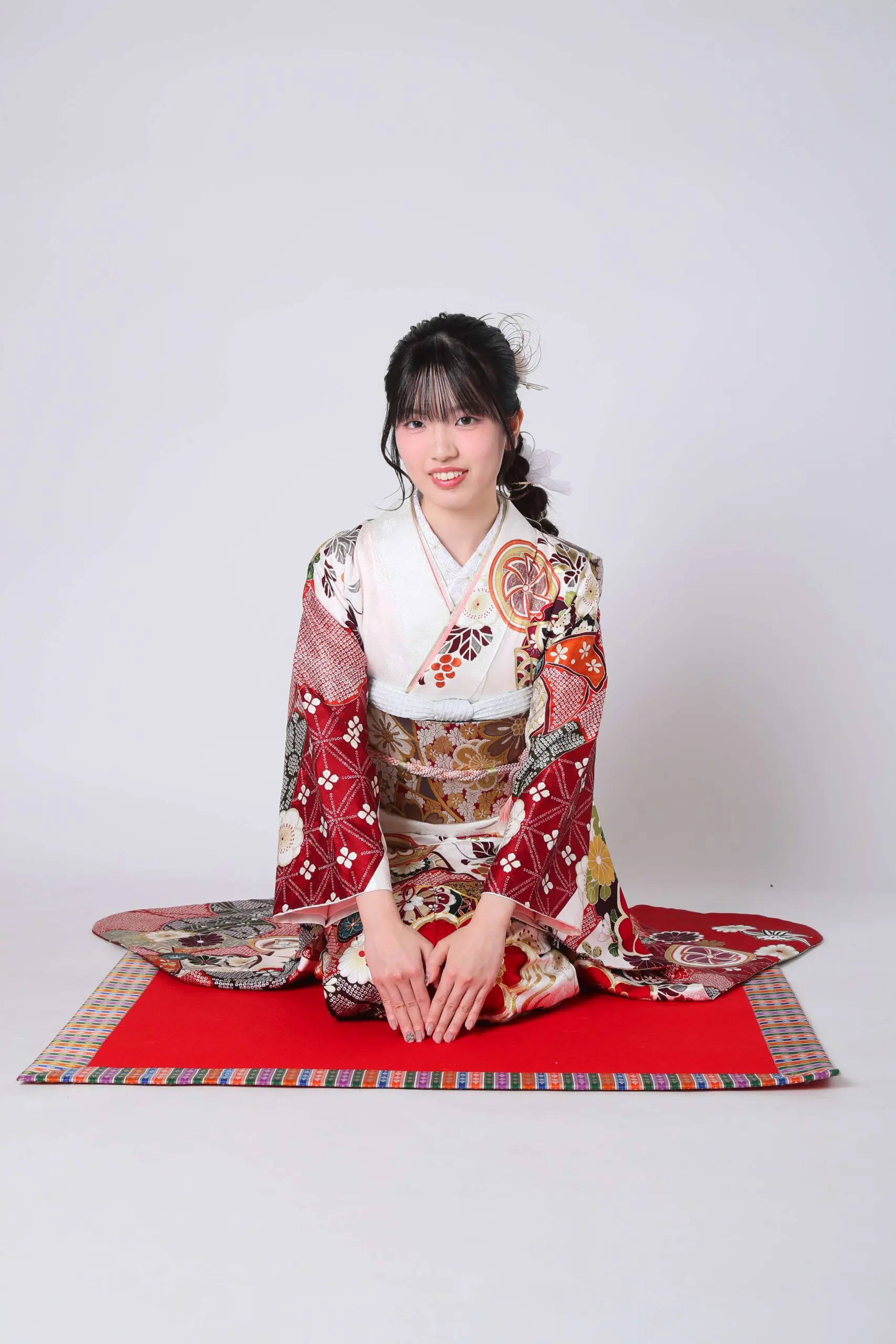 赤いマットの上に、赤、白、金の繊細な模様があしらわれた色鮮やかな着物を着た人物が座っている。背景はライトグレー。黒髪の人物は、令和7年成人式、戸田市のイベントでカメラに向かって優しく微笑んでいる。.