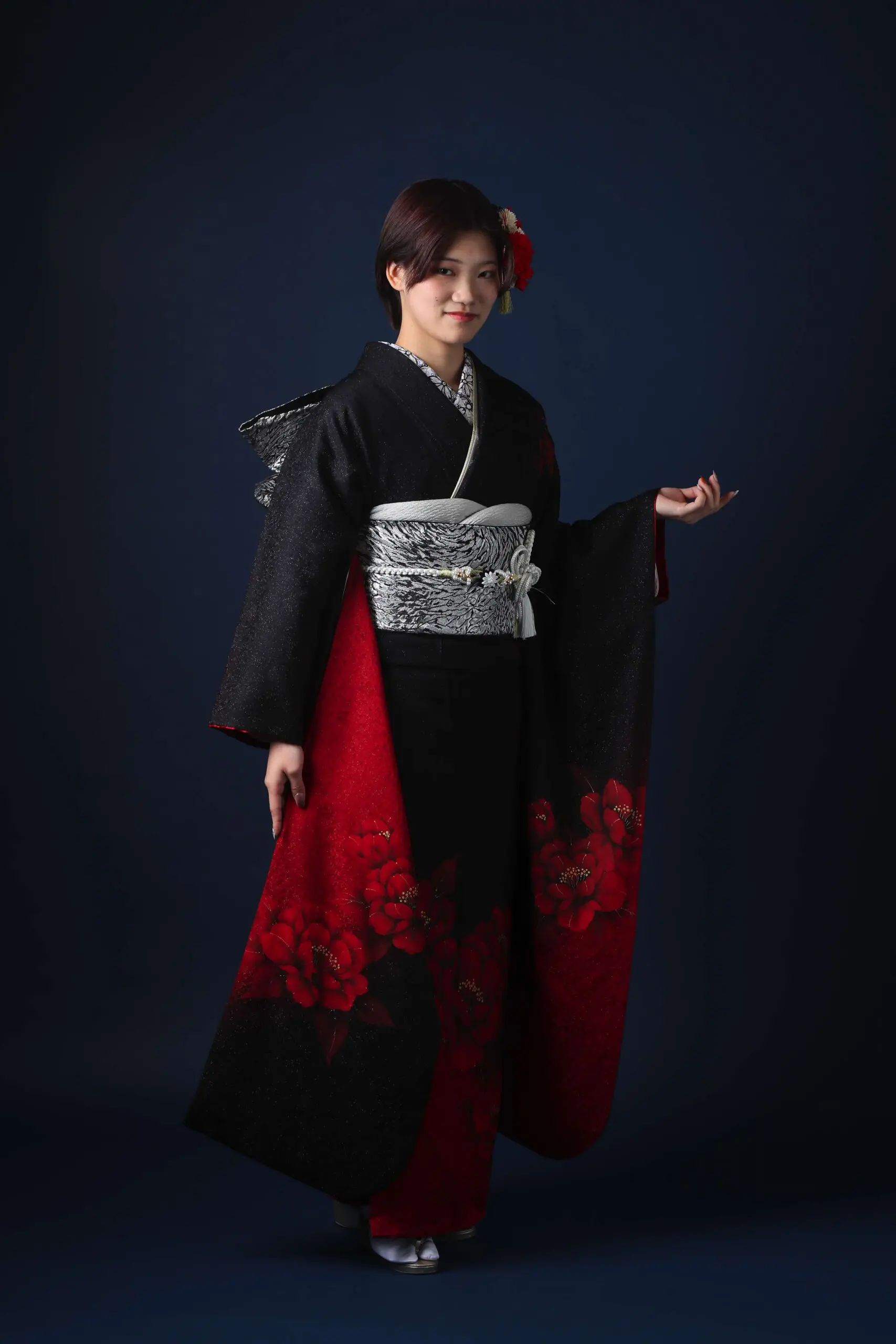 令和7年成人式、戸田市の着物を着ています。黒と赤の花柄と銀と白の帯、暗い背景に赤い花の髪飾りが特徴です。.