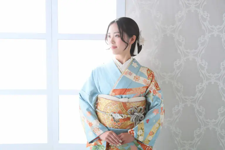 青、赤、金の複雑な模様が入ったカラフルな令和7年成人式、川口市の着物を着た人が室内の窓際に立っています。彼らの髪は、模様のある白い壁を背景に、白い花のアクセサリーで飾られています。.