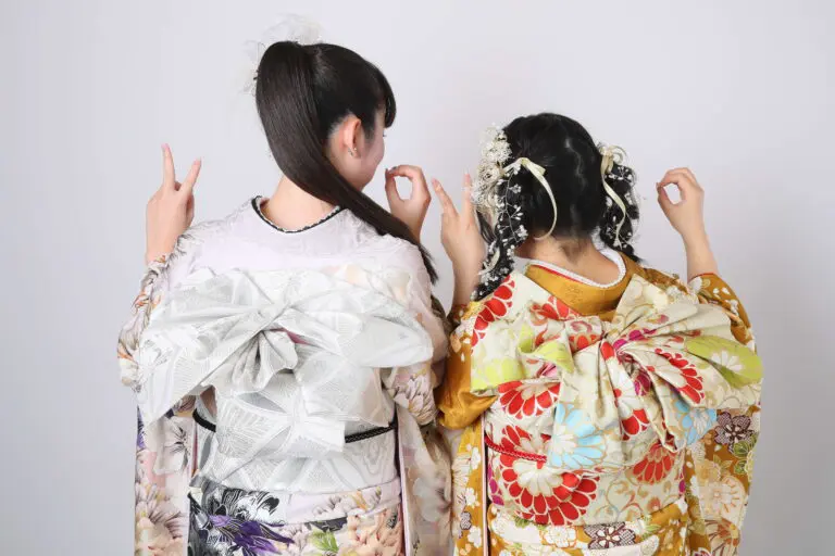 二人の女性が、複雑な帯結びが施された色鮮やかな着物を着て後ろ姿で写っている。二人とも髪を飾りで飾り、白無地の背景にピースサインをしながらポーズをとっている。.