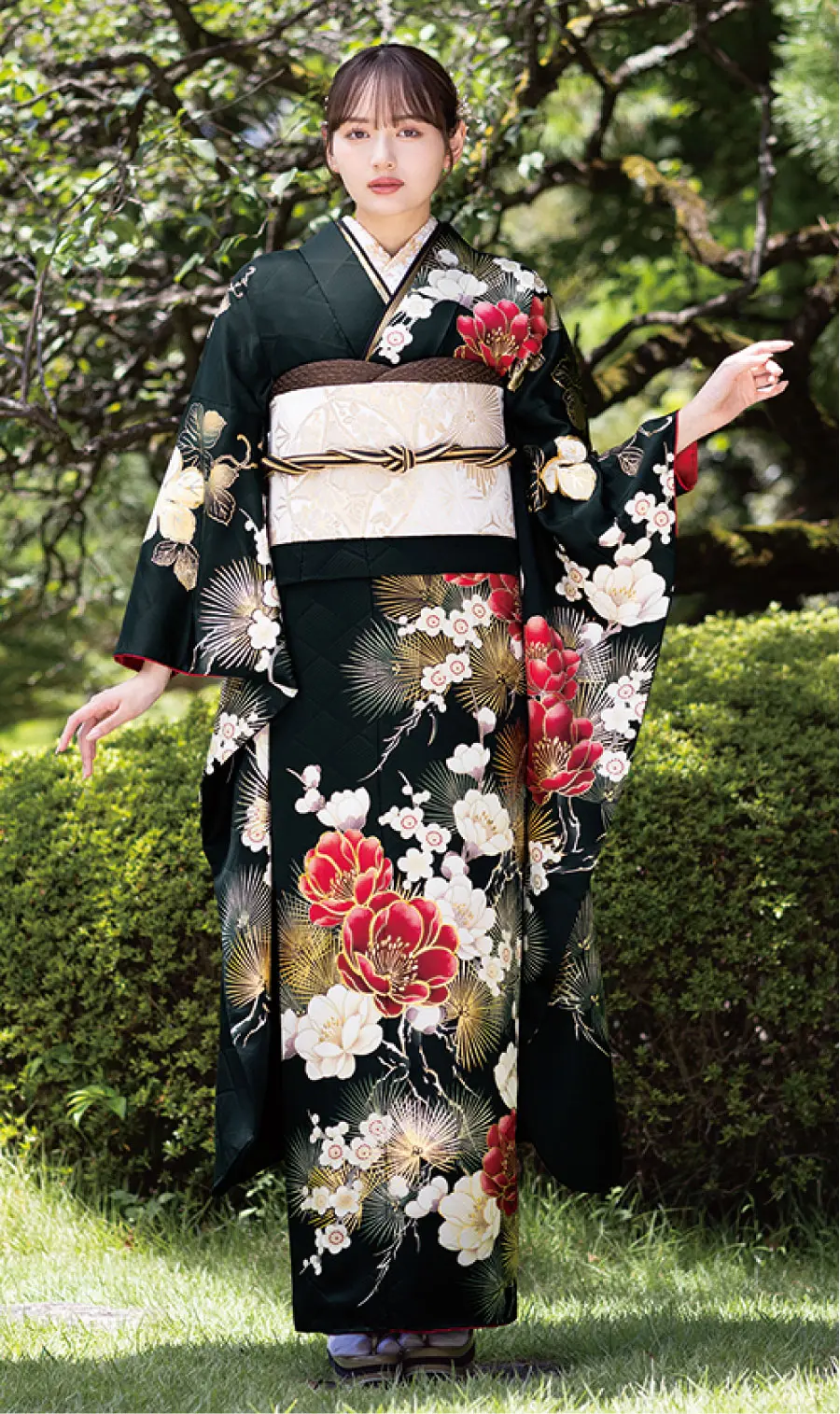 赤、白、金の花柄があしらわれたK109の黒い着物に、淡い金色の帯と草履を合わせた女性が屋外に立っている。緑と木々に囲まれ、左腕を軽く上げている。.