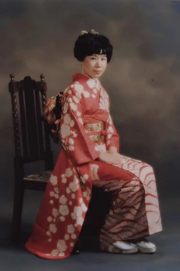 スタジオのシンプルな背景を背に、ピンクと赤の伝統的な花柄の着物、装飾的な帯、白い靴下とサンダルを身につけた女性が木製の椅子に座っています。.