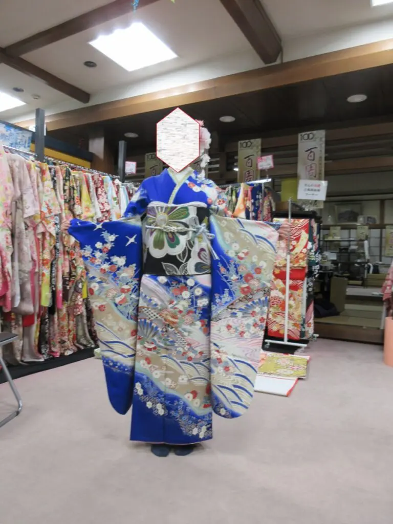 室内に、青い、精巧な模様の着物を着て、袖を広く伸ばした人物が立っています。おそらく振袖展の会場のようです。背景には色とりどりの着物が掛けられていますが、人物の顔は見えません。.