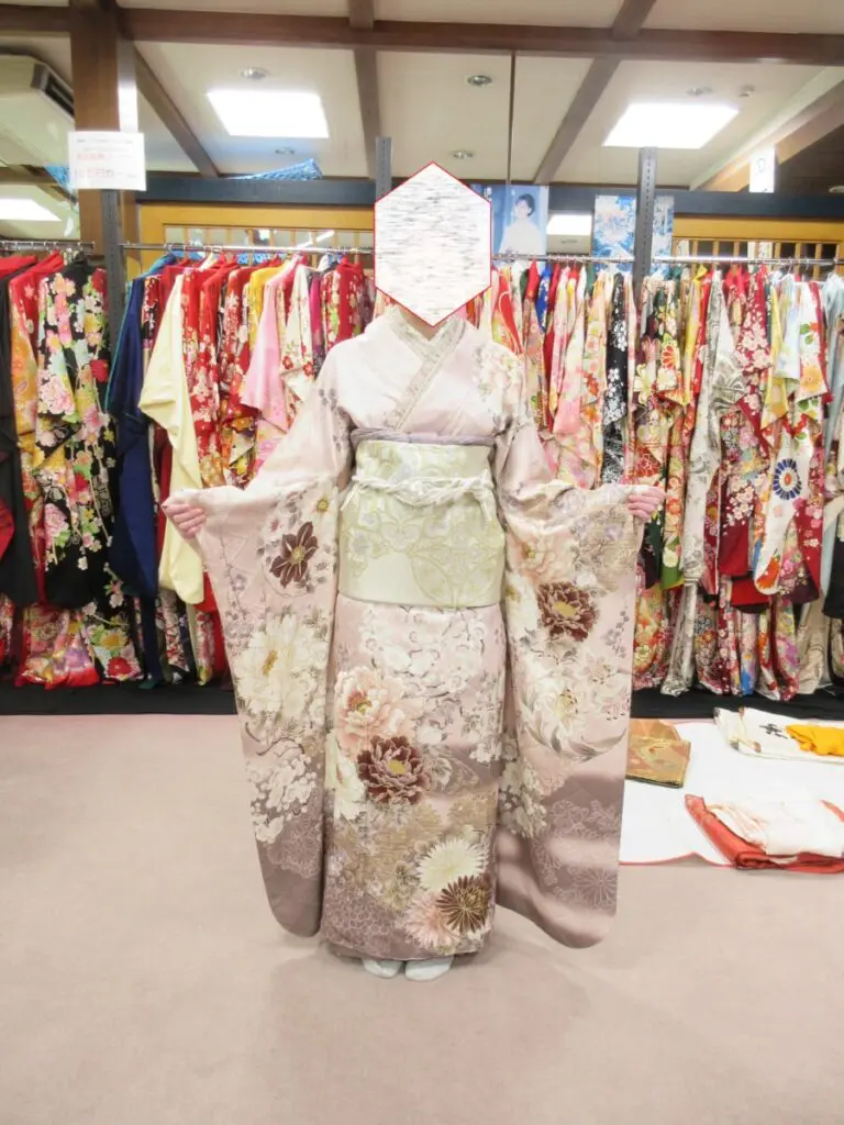 淡い色の花柄の着物を着た人物が、室内で腕を軽く伸ばして振袖展をしています。背景には色とりどりの着物がずらりと並び、人物の顔は見えません。.