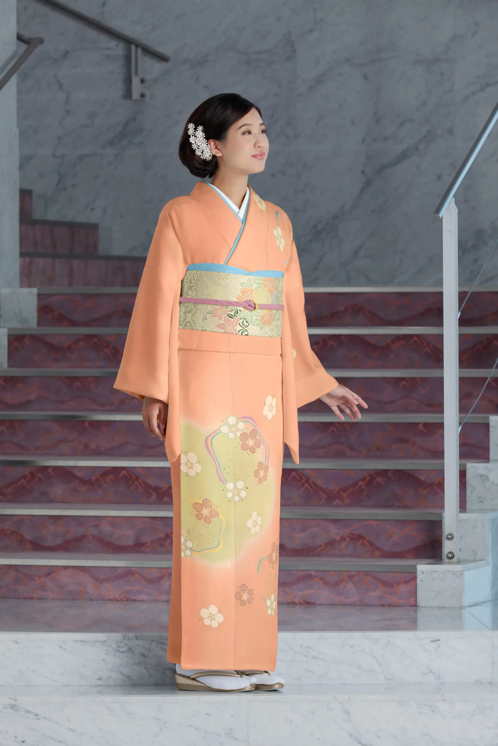 訪問着No.136 Mサイズ（桃色、花柄）を着た女性が大理石の階段の上に立ち、髪に花のアクセサリーをつけ、両手をリラックスさせたまま少し横を向いている。.