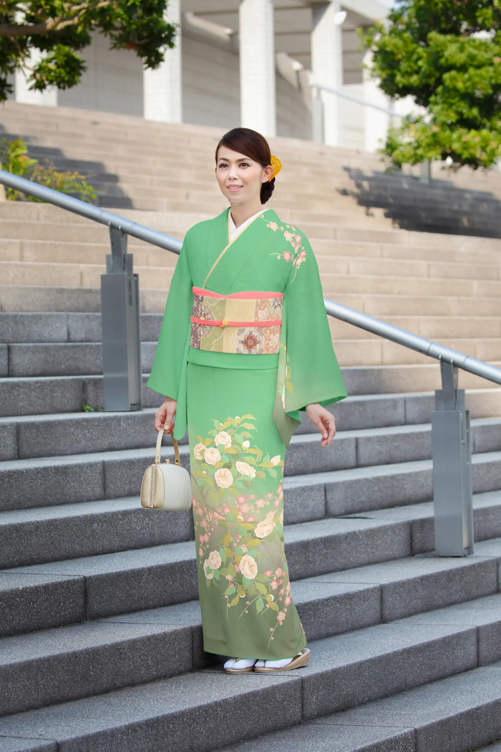 緑色の花柄の着物、訪問着No.138 Lサイズに身を包んだ女性が、小さなベージュのハンドバッグを持ちながら、緑と建物を背に屋外の石段に立っている。.