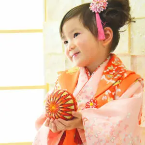 ピンクとオレンジ色の伝統的な着物と髪飾りを身に着けた幼い子供が、柔らかく照らされた室内の背景を背に、色鮮やかな刺繍のボールを持ち、微笑んで横を向いています。.