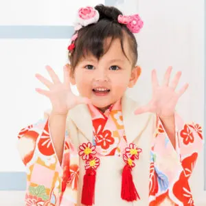 ピンクと赤の髪飾りを付けた色鮮やかな花柄の着物を着た若い女の子が、明るい背景の室内に立って、両手を前に広げて微笑んでいます。.