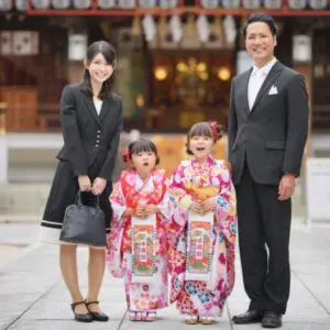 4人家族が屋外でポーズをとっています。色鮮やかな着物を着た2人の少女が、黒いフォーマルスーツを着た女性と男性の間に立っています。背景には伝統的な建物が見えます。.