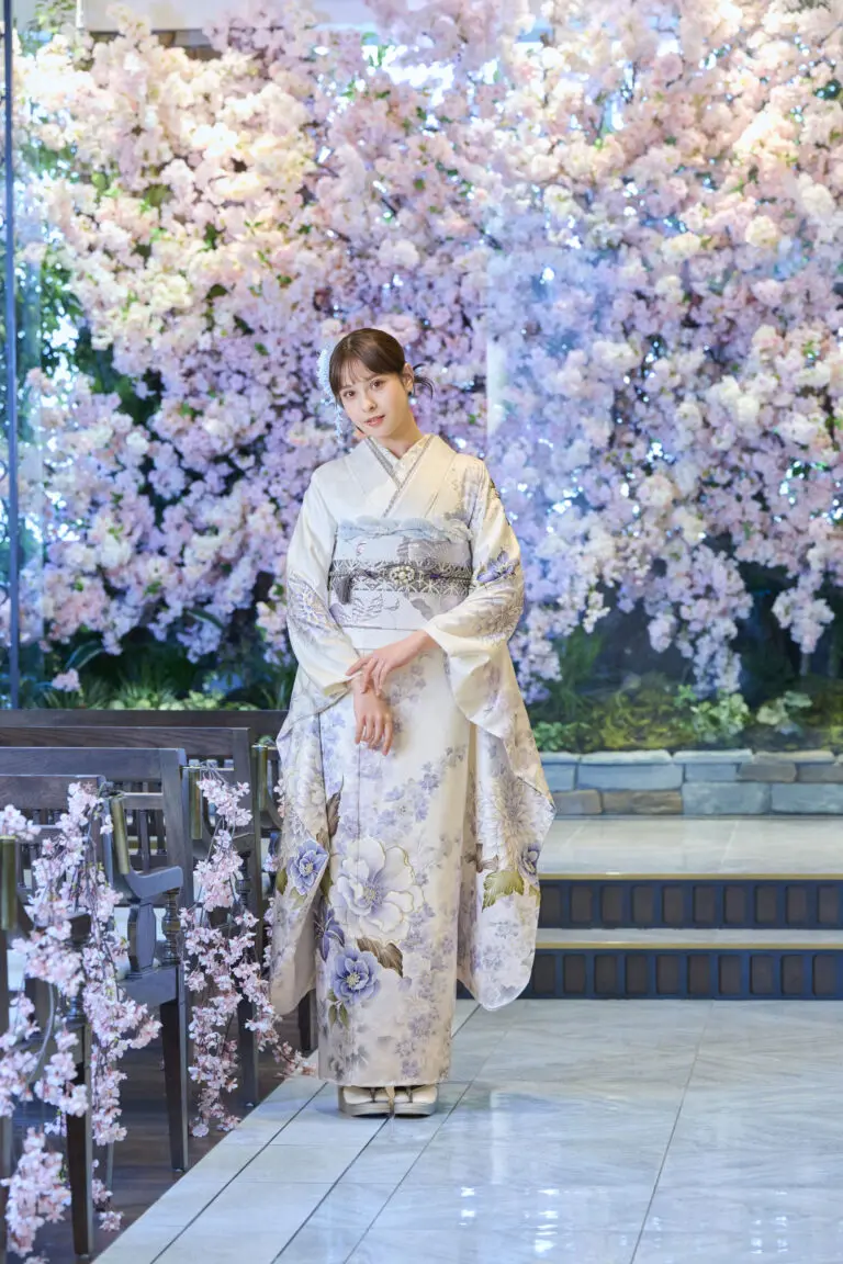 M-416の花模様の着物を着た女性が、室内に置かれた花で飾られたベンチの横に立っている。.