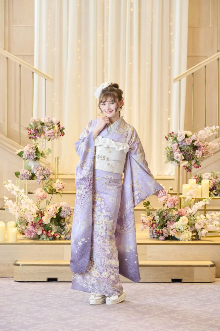 M-401のラベンダー色の着物に白い帯を締め、ピンク、白、クリーム色の花々に囲まれた明るい台の上でポーズをとる女性。.