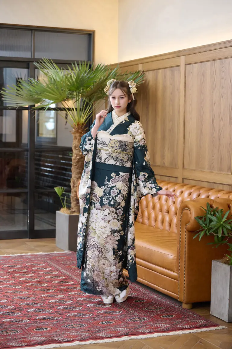 M-413のダークグリーンとクリーム色の花柄の着物を着た女性が、日焼けした革張りのソファ、木製の壁パネル、鉢植え、ヤシの木のそばの赤い柄のラグの上に立っている。.