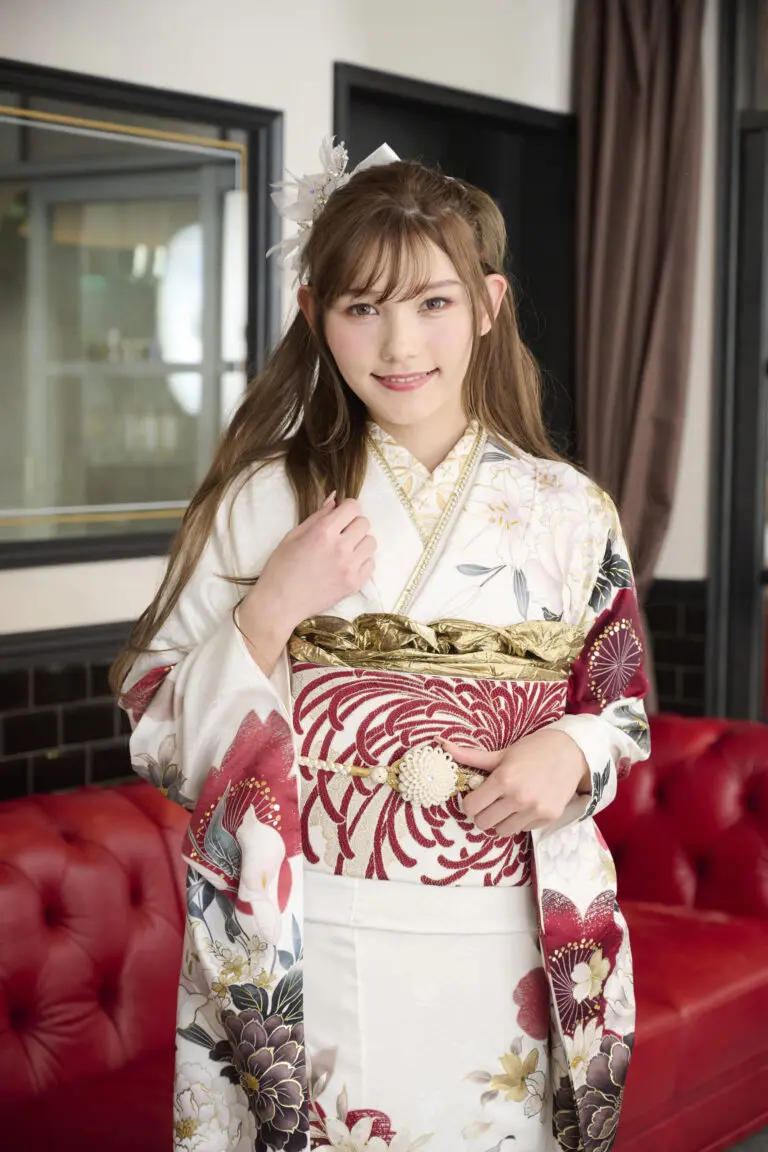 ライトブラウンのロングヘアの若い女性が、M-403の白と赤の花柄にゴールドのディテールが施された着物の帯を持ち、微笑みながら室内に立っている。背景には赤いソファと額縁入りの鏡が見える。.