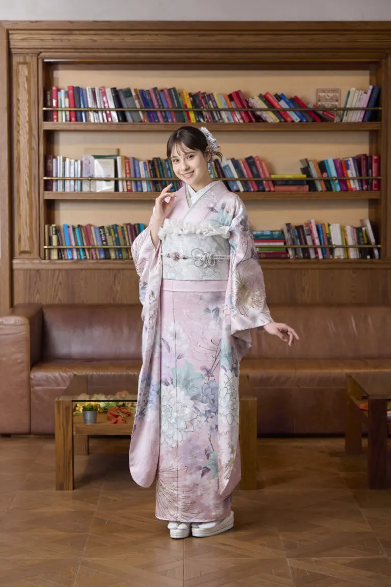 M-415の薄ピンクの花柄の着物を着た女性が本棚の前でポーズをとっている。片手は顔の近く、もう片手は脇に添えられている。背景には革張りのソファと木製のテーブルが見える。.