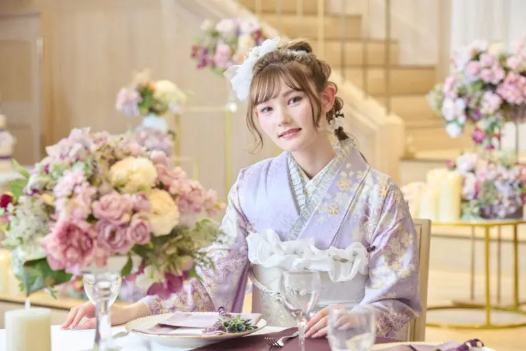 M-401の薄紫の花柄の着物を着た若い女性が、花、ガラス器、皿が置かれたテーブルに座っている。彼女がカメラを見つめているとき、背景には階段と追加の花飾りが現れる。.