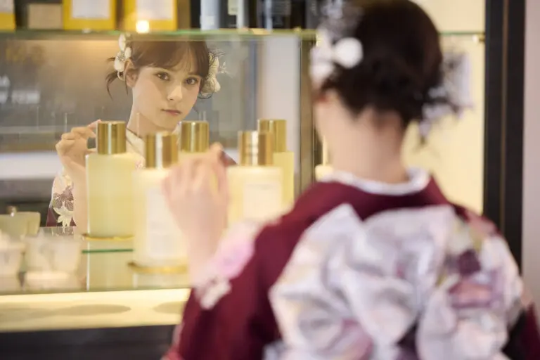 大きなリボンのついたマルーン色の着物を着た女性が、鏡に映った自分の姿を見ながら、棚に並んだ同じようなボトルの中からM-421のボトルに手を伸ばしている。.