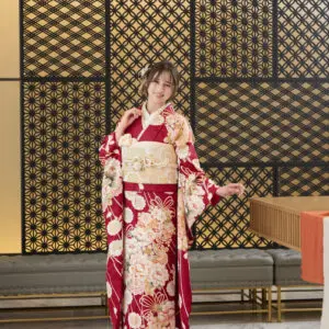 M-414の赤い花柄の着物にクリーム色の帯を締め、笑顔で片手を上げてポーズをとる人物が室内に立っている。背景は幾何学模様の装飾パネルと大理石の床。.