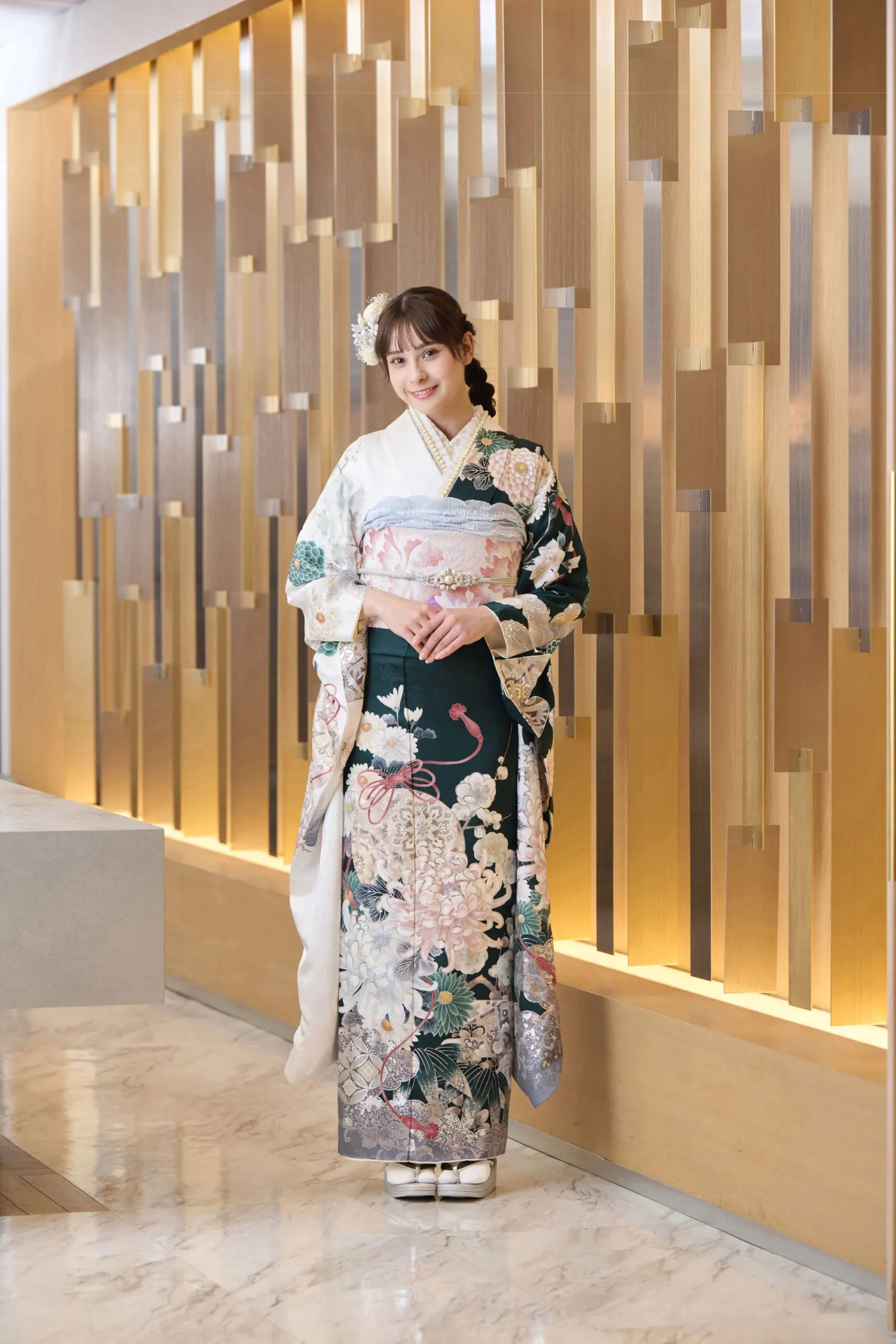 花模様のM-420の着物を着て、装飾的な髪飾りをつけた女性が、金色の幾何学模様のモダンな壁と磨き上げられた大理石の床の前に立っている。.