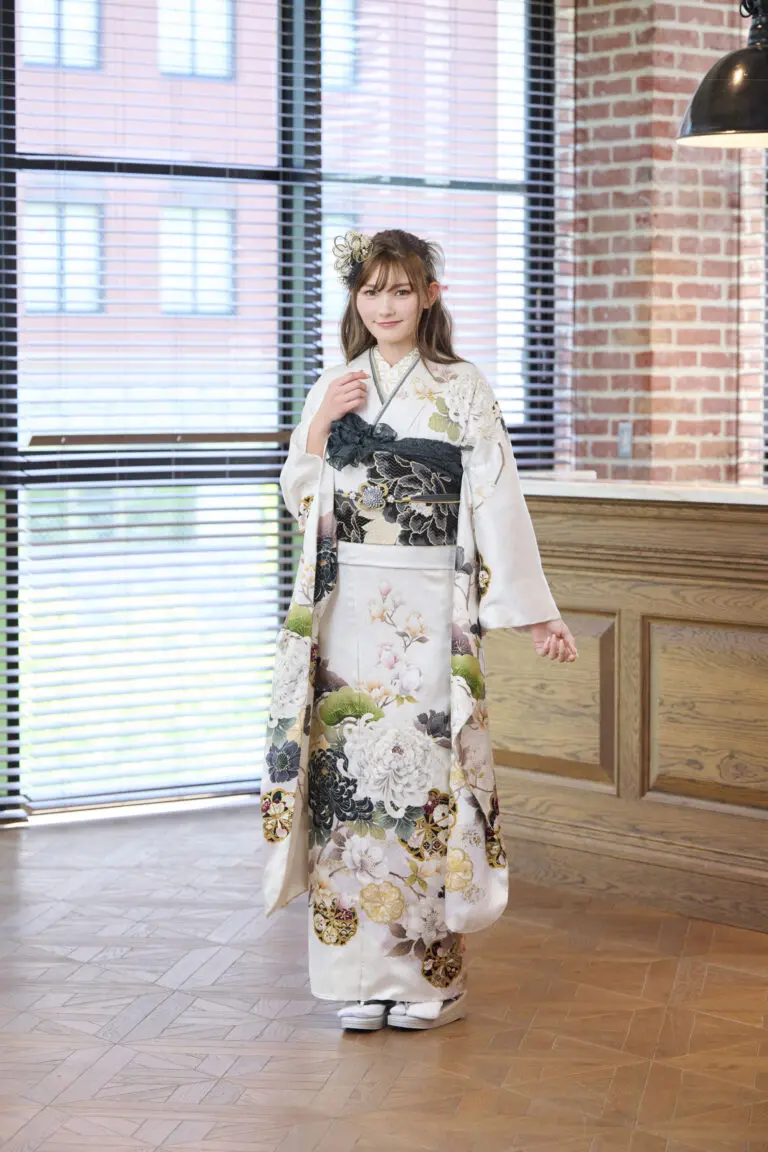 M-407の白い花柄の着物を着て、濃い色の帯を締めた、明るい茶色の髪を髪飾りでスタイリングした若い女性が室内に立っている。部屋はレンガの壁、大きな窓、フローリング。.