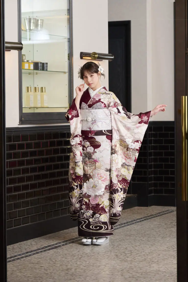 白、ワインレッド、ゴールドの花柄が特徴的なM-421フォーマル着物を着て、室内でポーズをとる女性。髪飾りをつけ、片手で顔を触り、もう片方の手で着物の袖を持っている。.