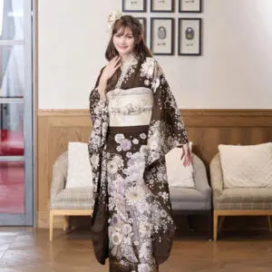 M-405の茶色の花柄の着物を着て、ベージュの帯を締めた女性が室内に立っている。髪に花を飾り、2つのソファの前でポーズをとり、後ろの壁には額装された肖像画が飾られている。.