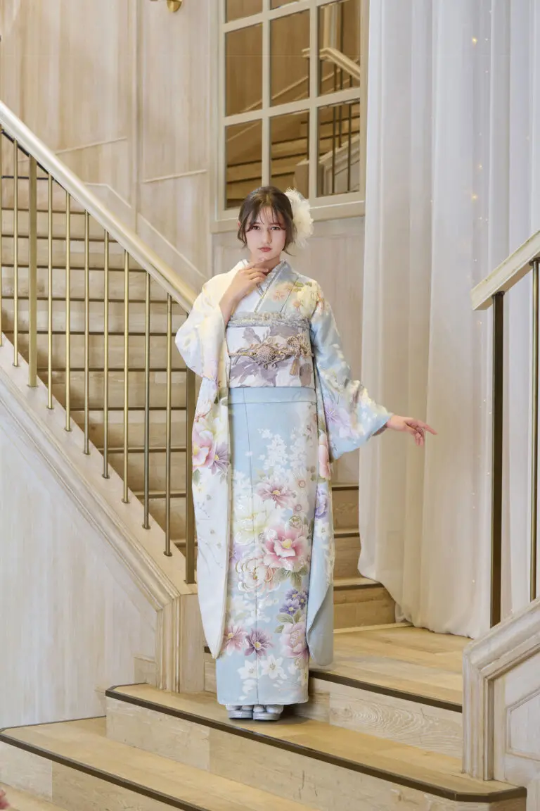 M-411の水色の花柄の着物を着た女性が、室内の木の階段に立っている。片手を顔の近くに置き、もう片方の腕を伸ばし、髪には大きな白い花のアクセサリーをつけている。.