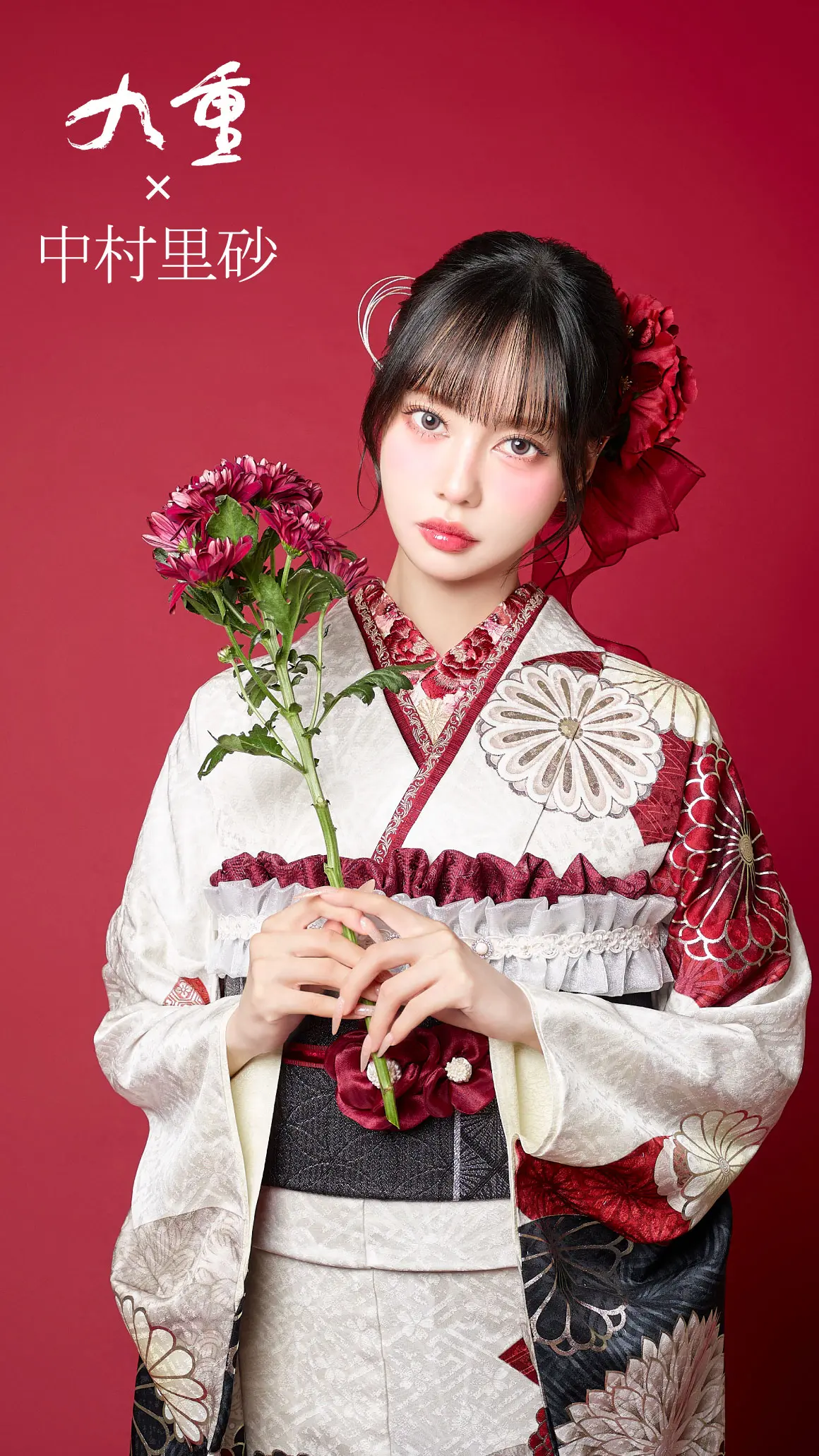柄物の着物を着た若い女性が、赤無地の背景の前で濃い赤色の花を持っている。前髪のあるストレートの黒髪で、花のヘアアクセサリーをつけている。左上に日本語の文字が見える。.
