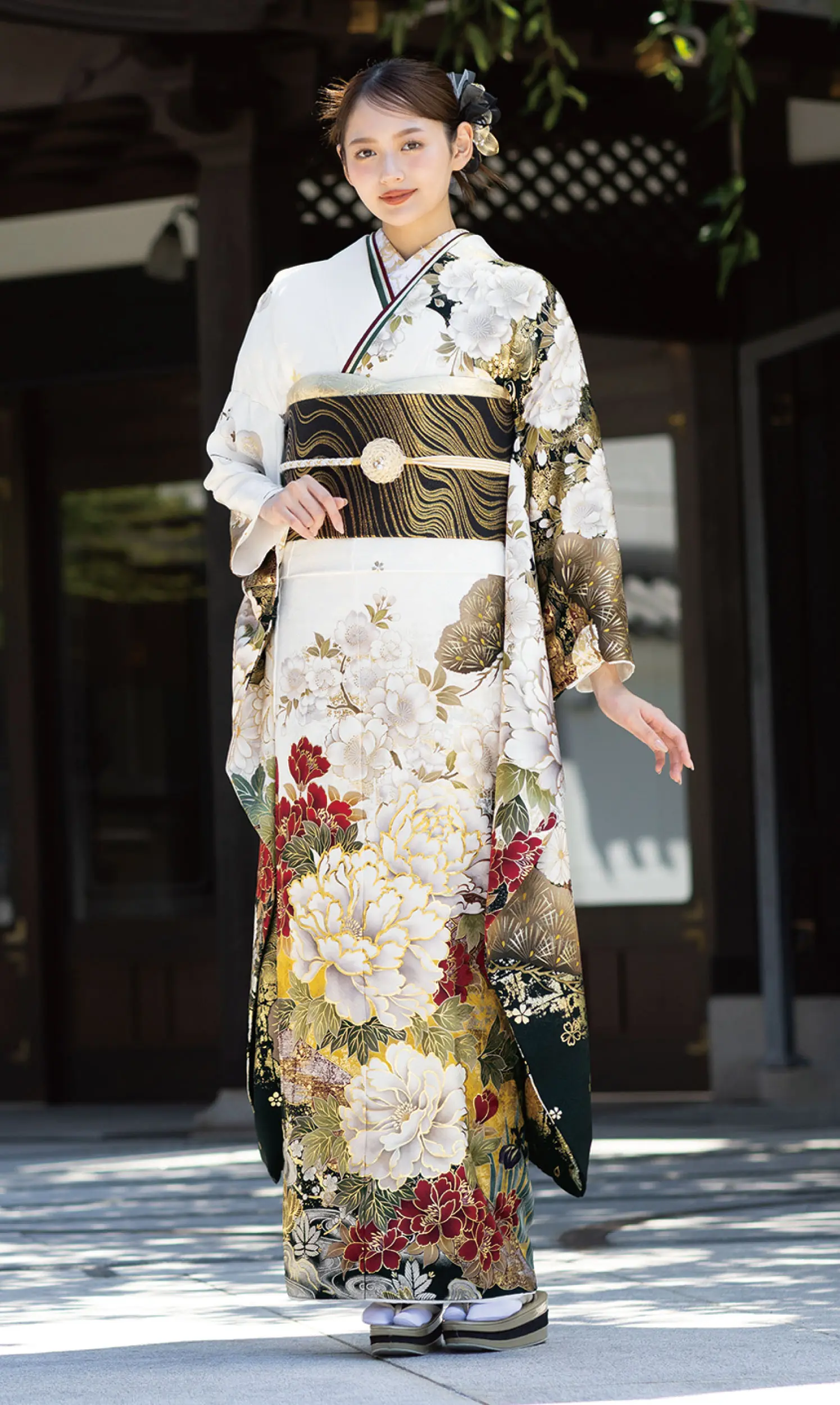 白、金、赤、緑の花柄に金と黒の縞の帯を合わせたK101フォーマル着物を着て、屋外でポーズをとる女性。髪に花を飾り、片手を少し上げている。.
