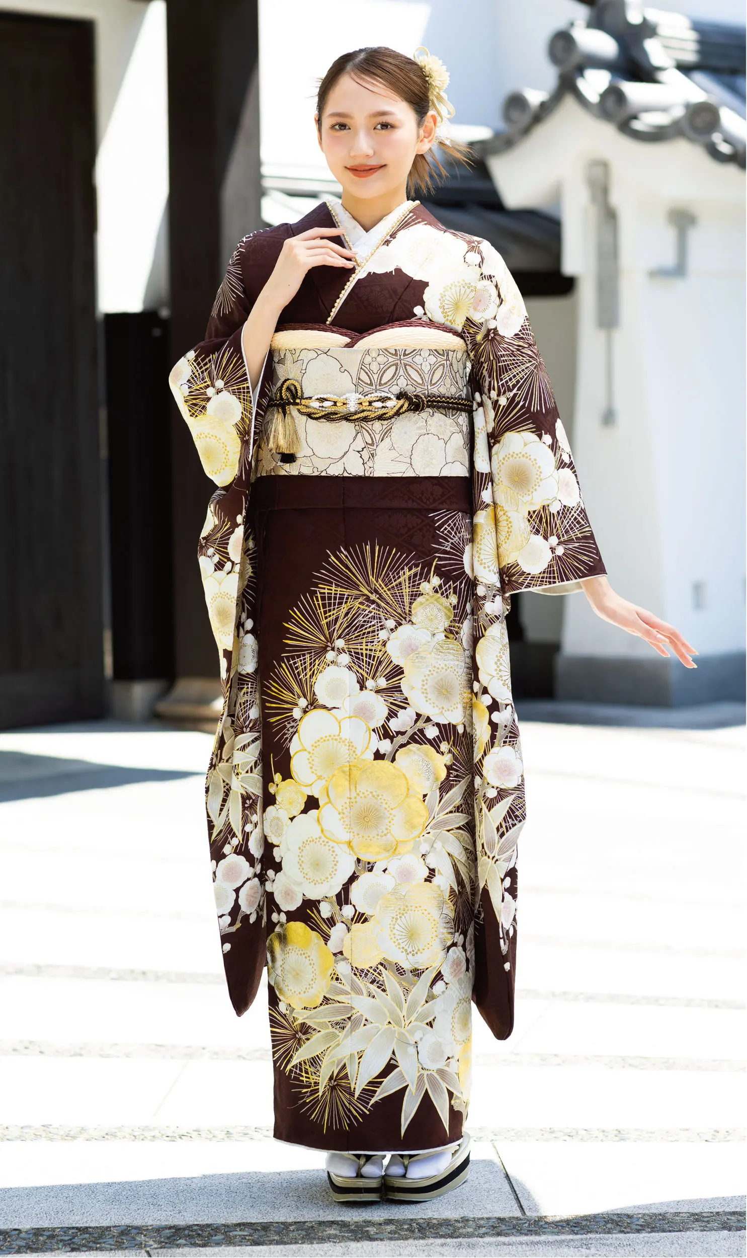 K101のこげ茶に黄色と白の花模様の着物を着て、ベージュの幅広の帯ベルトを締め、伝統的な草履を履いた女性が屋外に立っている。片手を上げてポーズをとり、優しく微笑んでいる。.
