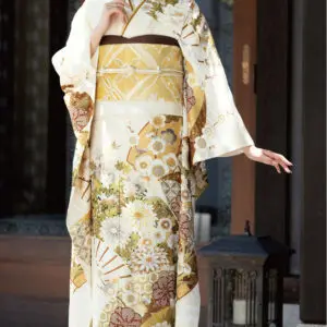 花柄や幾何学模様の入ったクリーム色と金色の伝統的な衣装、K101の着物を着て屋外でポーズをとる女性。花柄のヘアアクセサリーをつけ、木造の建物の近くで黒い提灯のそばに立っている。.