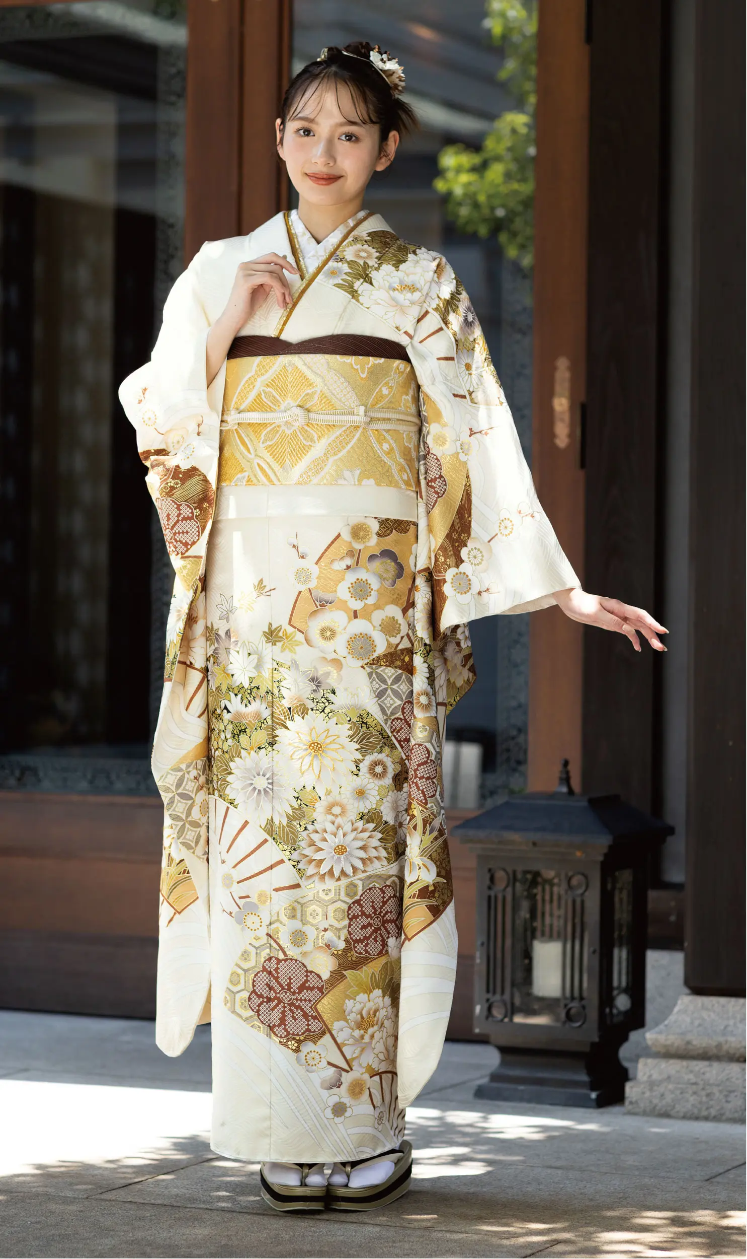 花柄や幾何学模様の入ったクリーム色と金色の伝統的な衣装、K101の着物を着て屋外でポーズをとる女性。花柄のヘアアクセサリーをつけ、木造の建物の近くで黒い提灯のそばに立っている。.