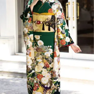 ダークグリーンとゴールドの花柄プリントのK101着物にゴールドの帯を締め、ガラス戸の前に立つ女性。髪はかんざしでアップにし、伝統的な草履に白い靴下を合わせている。.