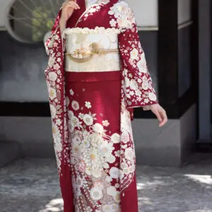 K101という伝統的な赤と白の花柄の着物に金の帯を締めた女性が屋外にいる。片手を顔の近くに優雅に上げ、横を見つめる彼女の髪を飾る花。.