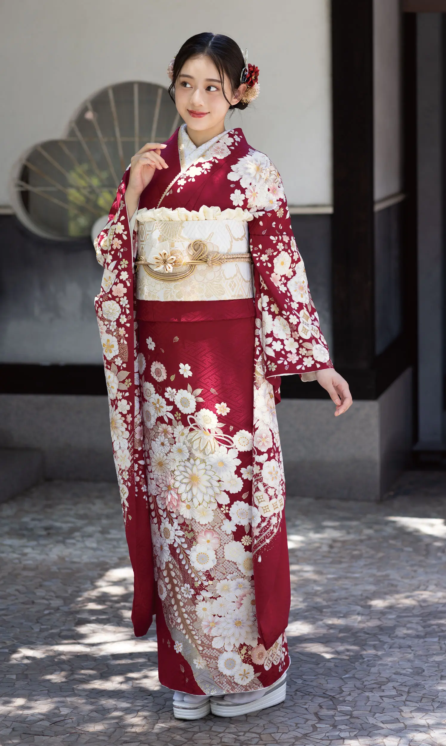 K101という伝統的な赤と白の花柄の着物に金の帯を締めた女性が屋外にいる。片手を顔の近くに優雅に上げ、横を見つめる彼女の髪を飾る花。.