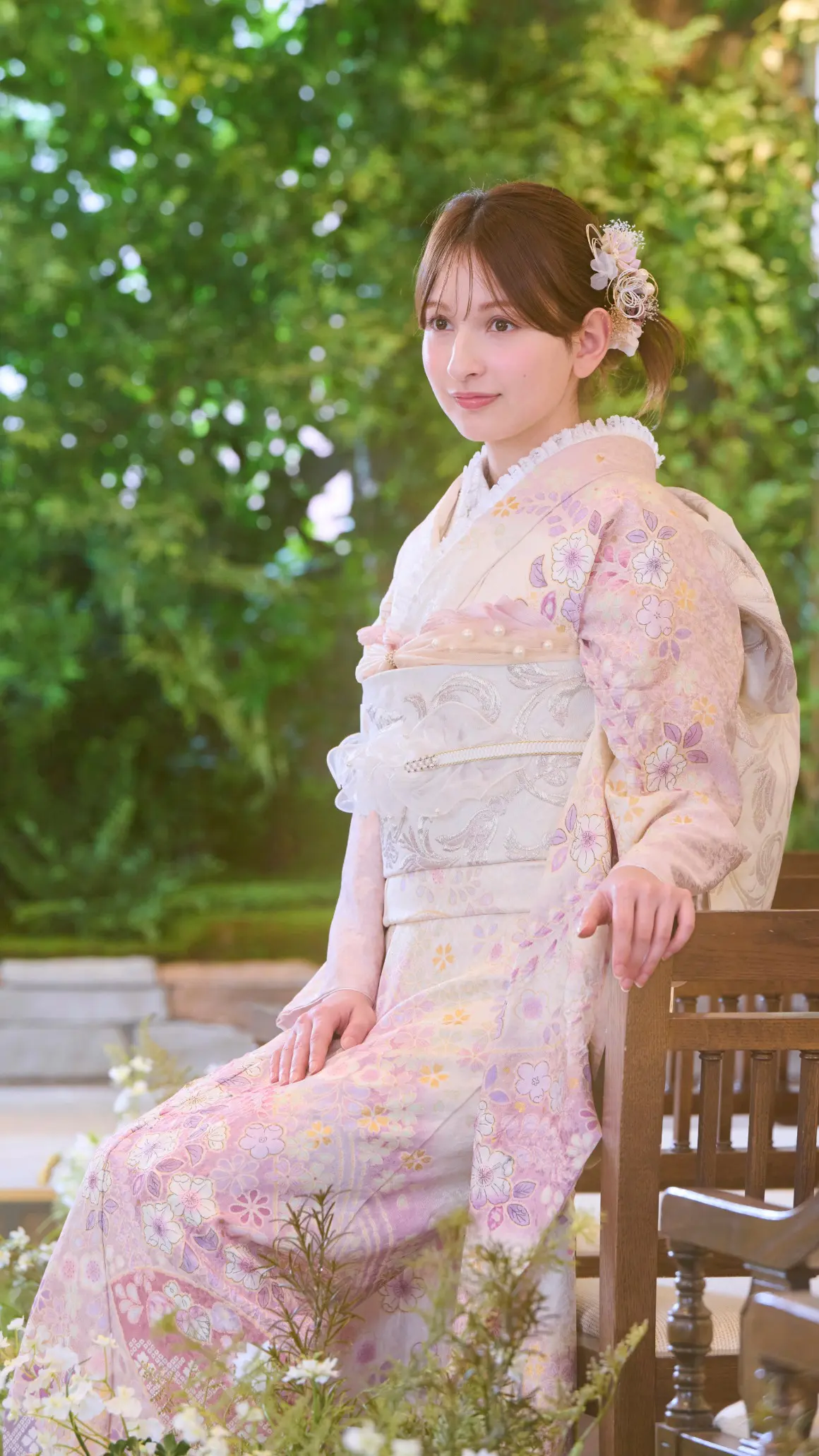 淡いピンク色の花柄の着物を着て、髪に装飾品をつけた人物が、緑を背景にした屋外の木製の椅子に座っている。.