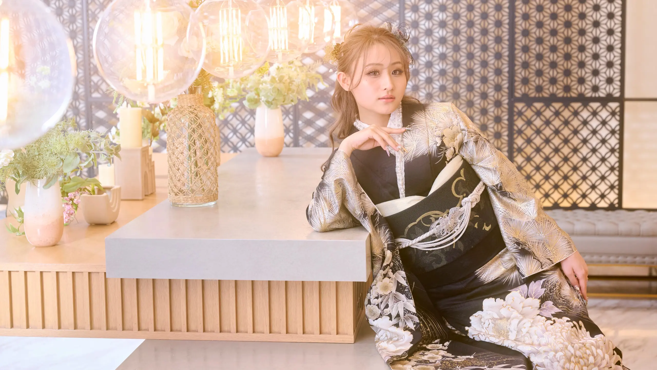 華やかな着物を着た女性が、モダンなカウンターに座り、肘をその上に載せている。背景には、装飾用の照明や観葉植物、精巧な間仕切りが見える。照明は温かみがあり、柔らかな光に包まれている。.